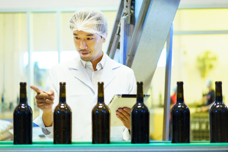 An empty glass bottles are rolling on the alcohol bottling belt machine in the manufacturing facility, meanwhile, male quality control supervisor is examining a bottles on the line.の写真素材
