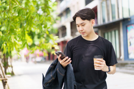Smart and good looking Asian young man in casual attire in the city or urban area with texting a message on smartphone while other hand holding a coffee cup. Man using smartphone to explore city map.の写真素材