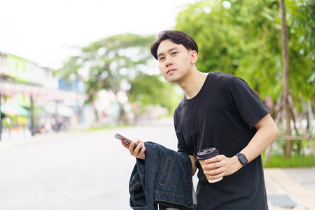 Smart and good looking Asian young man in casual attire in the city or urban area with texting a message on smartphone while other hand holding a coffee cup. Man using smartphone to explore city map.の写真素材