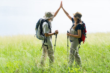 Man and woman in hiking outfits stand by a lake in a green field under soft morning sunlight,...
