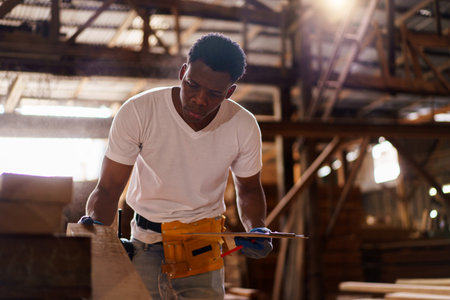 African carpenter works in a woodworking shop, using a tape measuring and pencil to mark on a wood...