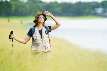 Happy Asian-Western woman in hiking outfit stands by a lake surrounded by green grass and soft morning sunlight, enjoying the peaceful beauty of nature and outdoor travel.の写真素材