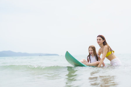 White Western woman and her young daughter enjoy surfing together on a sunny beach, smiling and laughing while spending quality time outdoors during their joyful summer vacation.の写真素材