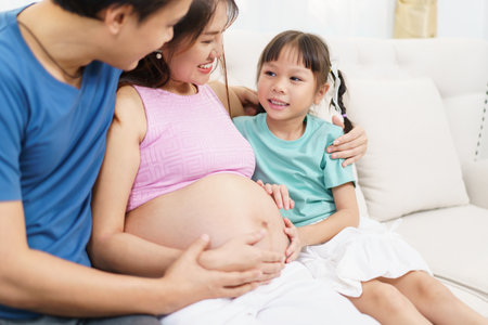 Asian family â husband, pregnant wife, and young daughter â relaxing and chatting together in a cozy living room. Daughter touches her mother's pregnant belly with excitement, love, and curiosity.の写真素材