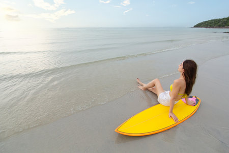 Western white woman in a bikini relaxing on the beach beside surfboard, enjoying peaceful moment while watching the sunset. Perfect seaside scene of freedom, and serenity. Summer activities concept.の写真素材