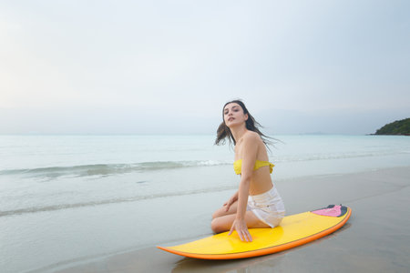 Western white woman in a bikini relaxing on the beach beside surfboard, enjoying peaceful moment while watching the sunset. Perfect seaside scene of freedom, and serenity. Summer activities concept.の写真素材