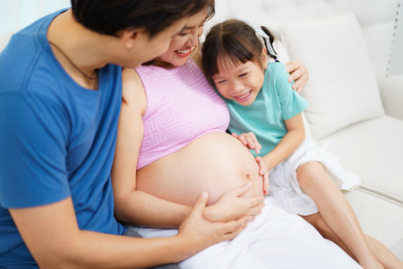 Asian family â husband, pregnant wife, and young daughter â relaxing and chatting together in a cozy living room. Daughter touches her mother's pregnant belly with excitement, love, and curiosity.の写真素材