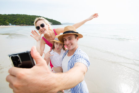 Happy caucasian white family including father, mother, and daughter are enjoy with taking a selfie photography on smartphone. A man using smartphone to take a selfie picture of a family at the beach.の写真素材