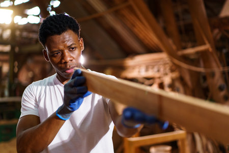African carpenter works in a woodworking shop, using a tape measuring and pencil to mark on a wood plank. This moment highlights craftsmanship, attention to detail, and professional woodworking skills.の写真素材