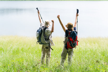 Man and woman in hiking outfits stand by a lake in a green field under soft morning sunlight, happily talking and enjoying the scenic nature view during their outdoor adventure.の写真素材