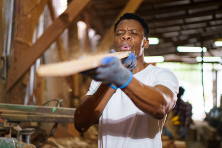 African carpenter works in a woodworking shop, using a tape measuring and pencil to mark on a wood plank. This moment highlights craftsmanship, attention to detail, and professional woodworking skills.の写真素材