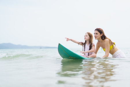 White Western woman and her young daughter enjoy surfing together on a sunny beach, smiling and laughing while spending quality time outdoors during their joyful summer vacation.の写真素材