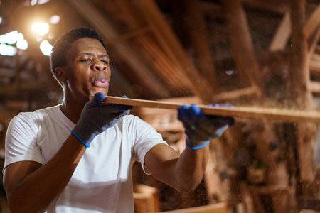 African carpenter works in a woodworking shop, using a tape measuring and pencil to mark on a wood plank. This moment highlights craftsmanship, attention to detail, and professional woodworking skills.の写真素材