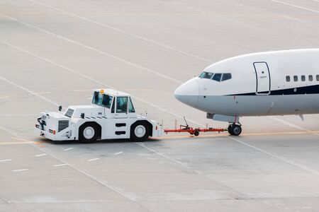 airplane on airport runway with pushback tractor attached to plane nose gear towing by ground vehicle to terminal gate.の写真素材