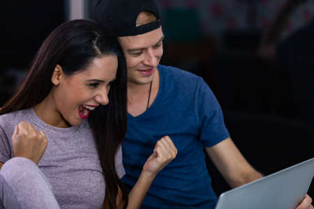 Happy young couple celebrate online victory, screaming with joy raising hands, looking at laptop screen together, wife and husband receive good news, achievement.の写真素材