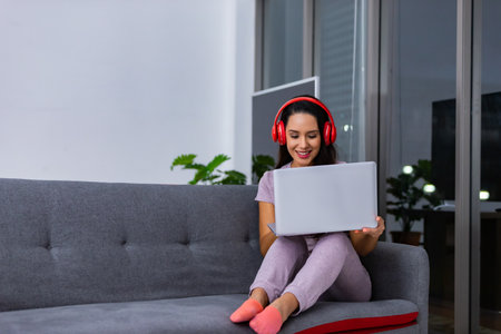 Happy women wearing headphones using a  laptop sitting on a couch in the night at homeの写真素材