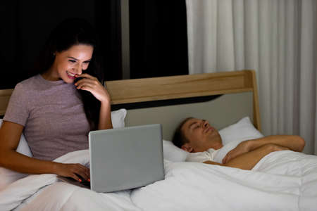 Woman sitting on bed in the night and using laptop computer while husband sleeping beside her.の写真素材