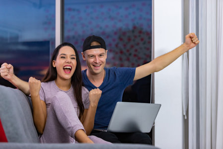 Happy young couple celebrate online victory, screaming with joy raising hands, looking at laptop screen together, wife and husband receive good news, achievement.の写真素材