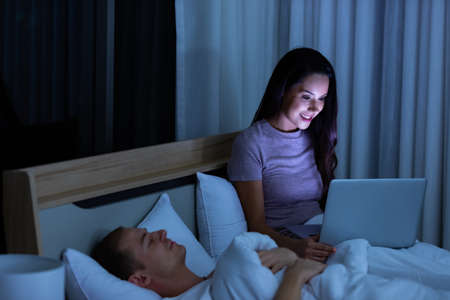 Woman sitting on bed in the night and using laptop computer while husband sleeping beside her.の写真素材