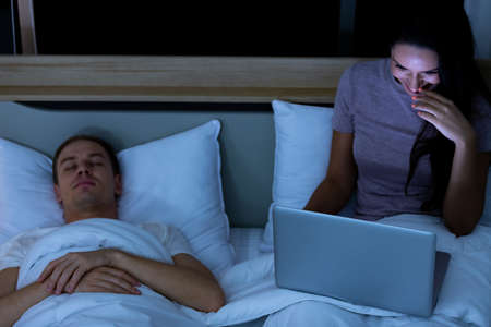 Woman sitting on bed in the night and using laptop computer while husband sleeping beside her.の写真素材