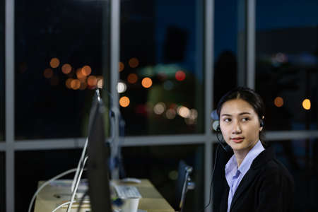 Asian young woman wearing headphone headset working overtime late at night in call center office. Concept for business at night.の写真素材