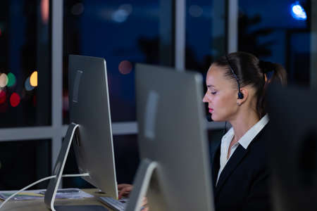 Caucasian young woman wearing headphone headset working overtime late at night in call center office with her colleague team. Concept for business at night.の写真素材
