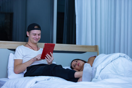 Man sitting on bed in the night and using tablet while wife sleeping beside him.の写真素材