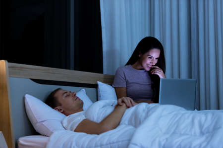 Woman sitting on bed in the night and using laptop computer while husband sleeping beside her.の写真素材