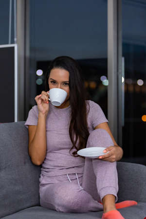 Woman enjoying a cup of tea in the night sitting on a couch in the living room at homeの写真素材