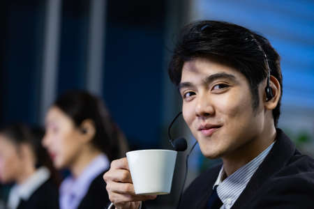 Businessman drinking coffee and wearing headphone headset working overtime late at night in call center office. Concept for business at night.の写真素材