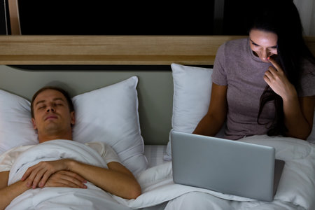 Woman sitting on bed in the night and using laptop computer while husband sleeping beside her.の写真素材