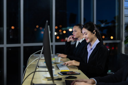 Asian young woman wearing headphone headset working overtime late at night in call center office with her colleague team. Concept for business at night.の写真素材