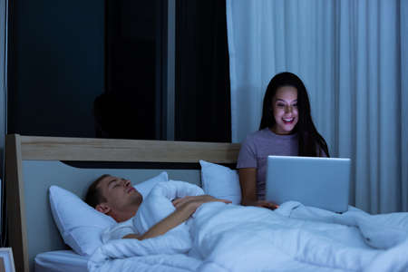 Woman sitting on bed in the night and using laptop computer while husband sleeping beside her.の写真素材