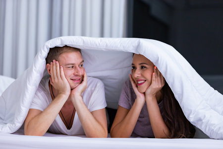 Portrait of young happy playful couple relaxing in comfortable cozy bed, cheerful smiling man and woman having fun posing covering head with soft warm blanket duvet on sleepoverの写真素材