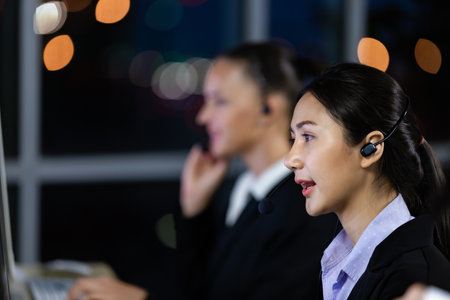 Asian young woman wearing headphone headset working overtime late at night in call center office with her colleague team. Concept for business at night.の写真素材