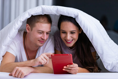 Portrait of young happy playful couple relaxing in comfortable cozy bed, cheerful smiling man and woman having fun posing covering head with soft warm blanket duvet on sleepoverの写真素材