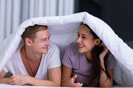 Portrait of young happy playful couple relaxing in comfortable cozy bed, cheerful smiling man and woman having fun posing covering head with soft warm blanket duvet on sleepoverの写真素材