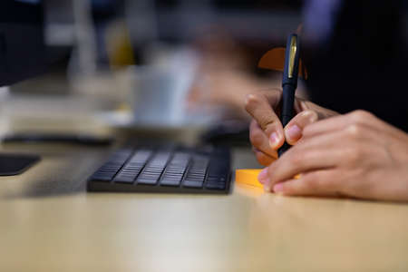 Close up image of businessman writing on an adhesive note at table in office at night, focus on hand.の写真素材