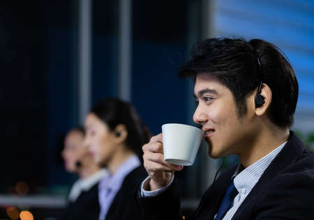 Businessman drinking coffee and wearing headphone headset working overtime late at night in call center office. Concept for business at night.の写真素材