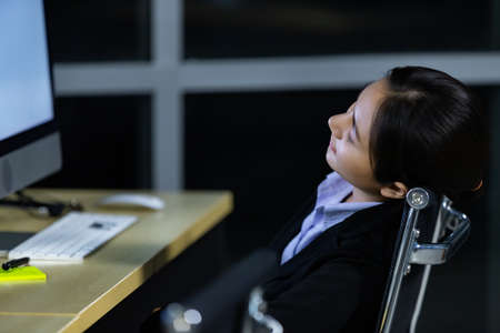 Asleep exhausted Asian lady call center worker employee sleeping on dest at night tired from late working alone in office. Ider for suffering and hard job of support team, light bokeh in background.の写真素材