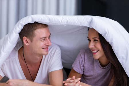 Portrait of young happy playful couple relaxing in comfortable cozy bed, cheerful smiling man and woman having fun posing covering head with soft warm blanket duvet on sleepoverの写真素材
