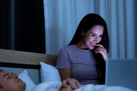 Woman sitting on bed in the night and using laptop computer while husband sleeping beside her.の写真素材