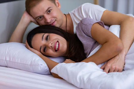 Couple lying comfortable sheets bed white blanket hugging joyful hold touch hands eyes closed wear pajama room indoorsの写真素材