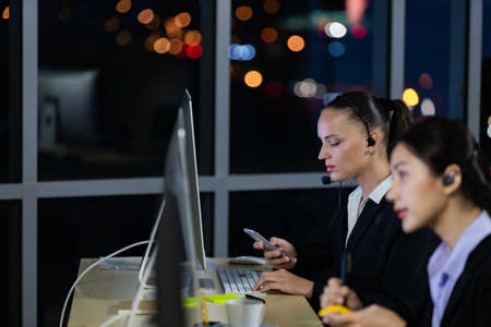 Caucasian young woman wearing headphone headset working overtime late at night in call center office with her colleague team. Concept for business at night.の写真素材