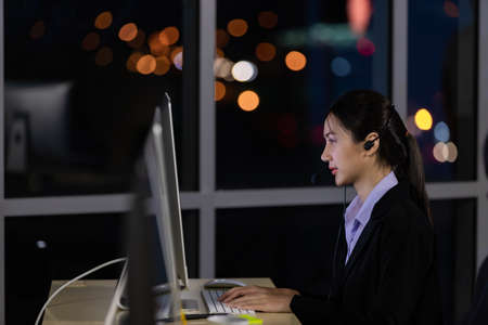 Asian young woman wearing headphone headset working overtime late at night in call center office. Concept for business at night.の写真素材