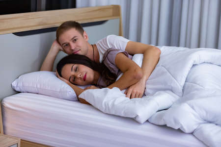 Couple lying comfortable sheets bed white blanket hugging joyful hold touch hands eyes closed wear pajama room indoorsの写真素材