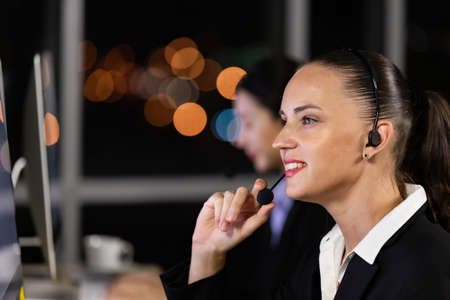 Caucasian young woman wearing headphone headset working overtime late at night in call center office with her colleague team. Concept for business at night.の写真素材