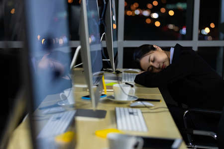 Asleep exhausted Asian lady call center worker employee sleeping on dest at night tired from late working alone in office. Ider for suffering and hard job of support team, light bokeh in background.の写真素材