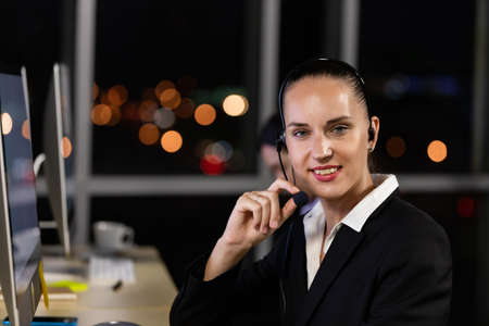 Caucasian young woman wearing headphone headset working overtime late at night in call center office with her colleague team. Concept for business at night.の写真素材