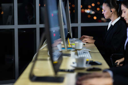 Caucasian young woman wearing headphone headset working overtime late at night in call center office with her colleague team. Concept for business at night.の写真素材
